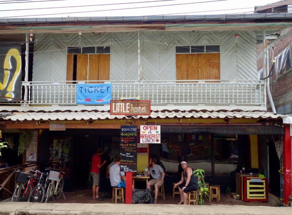 Outdoor seating area in front of a budget hostel: guests sit at tables under a corrugated roof, bicycles parked to the left, chalkboard menu nearby.