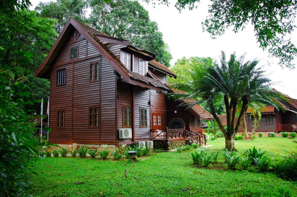 Wooden cabin with a brown exterior in a lush green garden, featuring a small veranda and palm trees nearby.