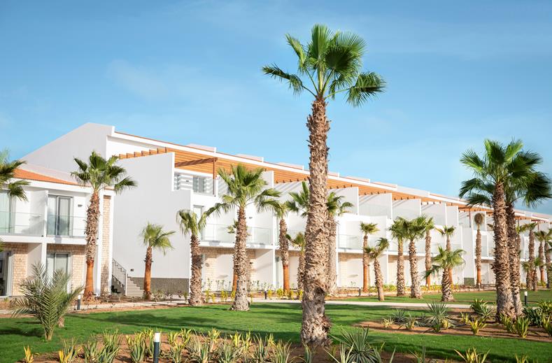 Row of white modern hotel villas with balconies, palm trees, and a green lawn under a blue sky.
