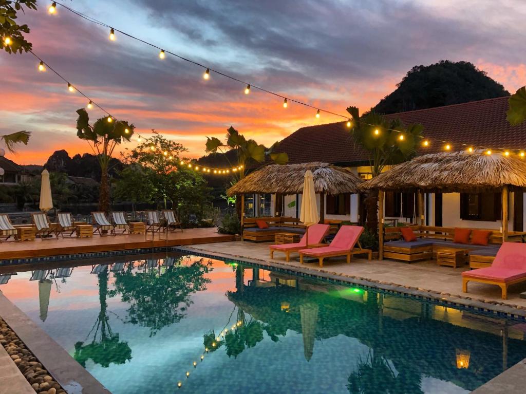 Poolside resort scene at sunset with string lights, palm trees and thatched cabanas.