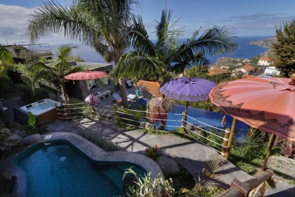 Terraced pool area with colorful umbrellas, palm trees, and a sea-view balcony overlooking a coastal town.