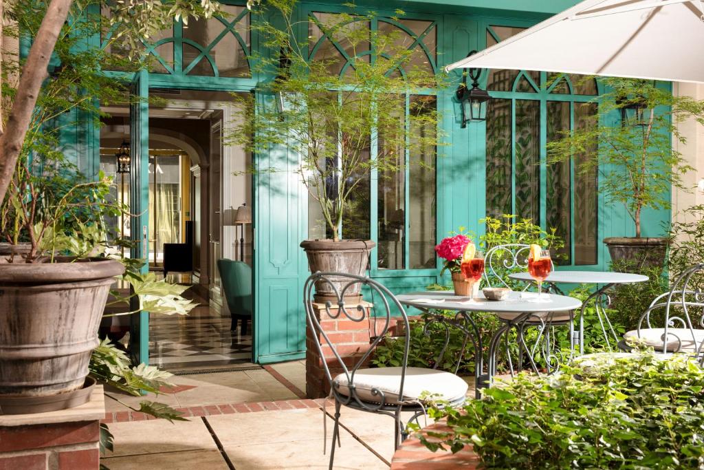 Outdoor cafe patio with teal doors, wrought-iron tables, potted trees, and two cocktails under a white umbrella on a sunny day.