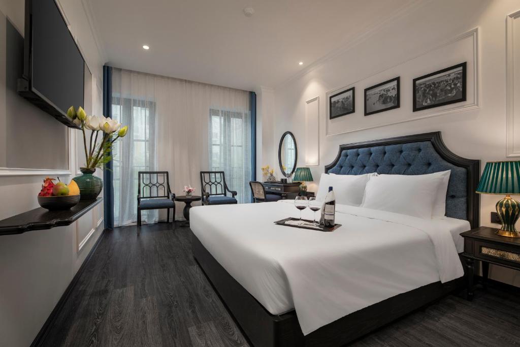 Luxurious hotel room with a large bed, tufted blue headboard, white linens, and a wine glass on a tray at the foot of the bed; green bedside lamps and dark wood furniture throughout.