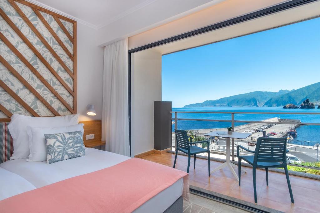 Bright hotel room with a bed, coral throw, palm-patterned pillow, and a balcony opening to blue sea views.