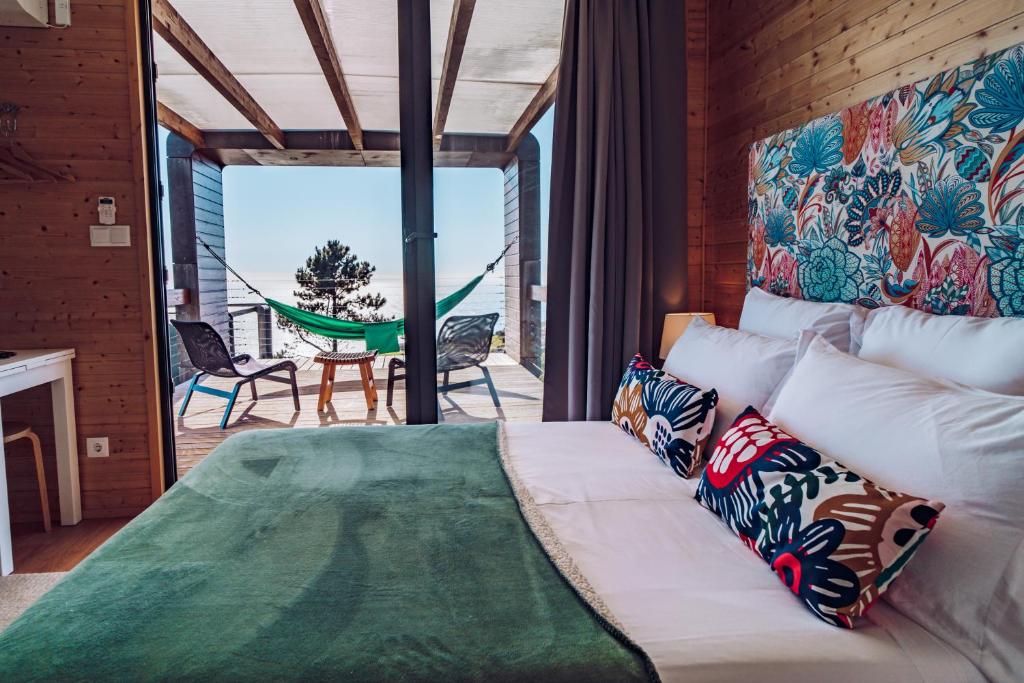 Cozy bedroom in a wooden cabin with a floral headboard, white pillows, and a green throw; sliding doors open to a deck with a hammock and ocean view.