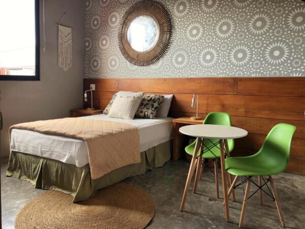 Cozy bedroom with a queen bed, beige quilt, pillows, two green mid-century chairs, and a round white table.
