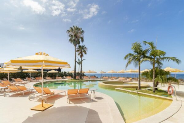 Sunlit resort pool area with yellow-striped umbrellas, orange lounge chairs, and palm trees against a blue sky near the sea.