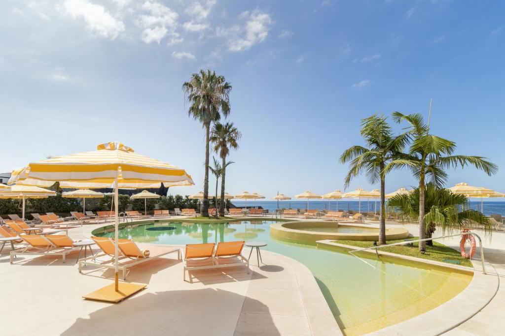 Sunlit resort pool area with yellow-striped umbrellas, orange lounge chairs, and palm trees against a blue sky near the sea.