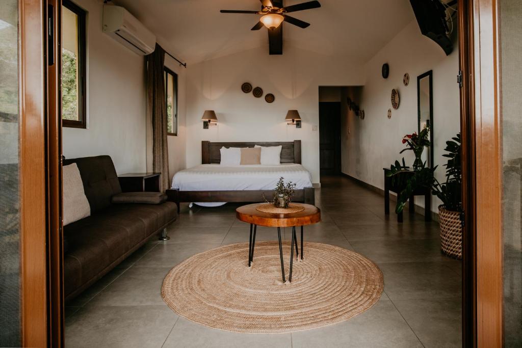 Open hotel room with a king bed at the far end, brown sofa on the left, round wooden coffee table, and plants nearby.