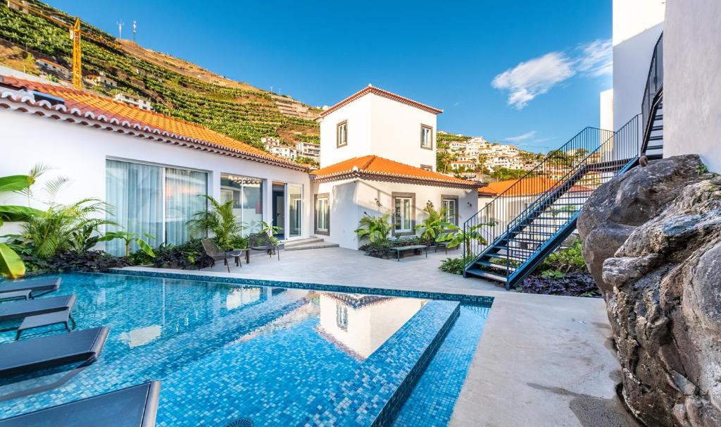 Villa courtyard with a rectangular blue pool, white walls, and hillside houses under a clear blue sky.