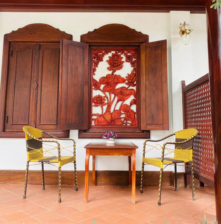 Two yellow wicker chairs sit on either side of a small wooden table with a vase of purple flowers, in front of carved red floral panels framed by dark wooden cabinets.