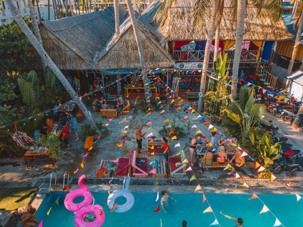Tropical poolside bar with thatched huts, palm trees, and colorful bunting; people relax and chat around tables.