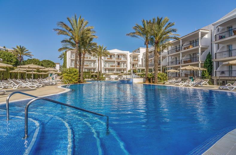 Resort pool area with palm trees, lounge chairs, and white buildings in the background.