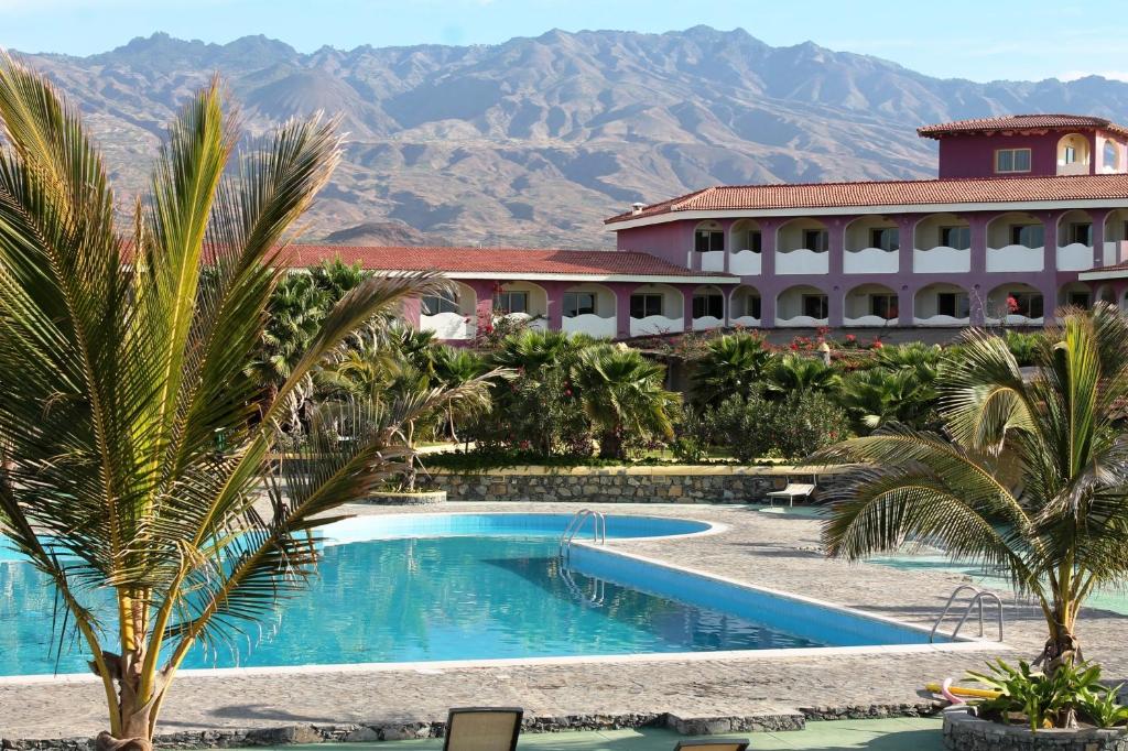 Resort pool area with palm trees, a pink arched building, and distant mountains behind the courtyard.