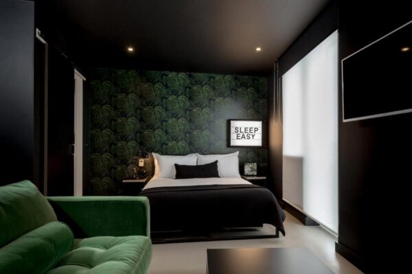 Hotel bedroom with a dark tropical wallpaper accent wall, a neatly made bed, and a green velvet sofa in the foreground.
