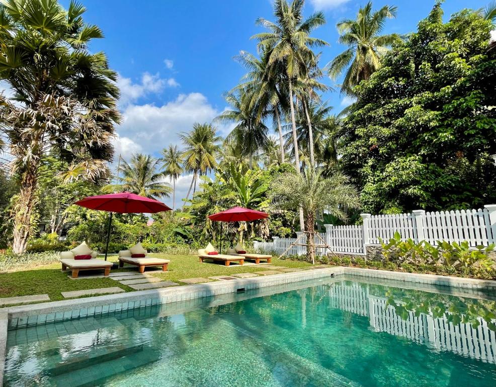 Poolside area with turquoise water, red umbrellas, and wooden lounge chairs under palm trees.