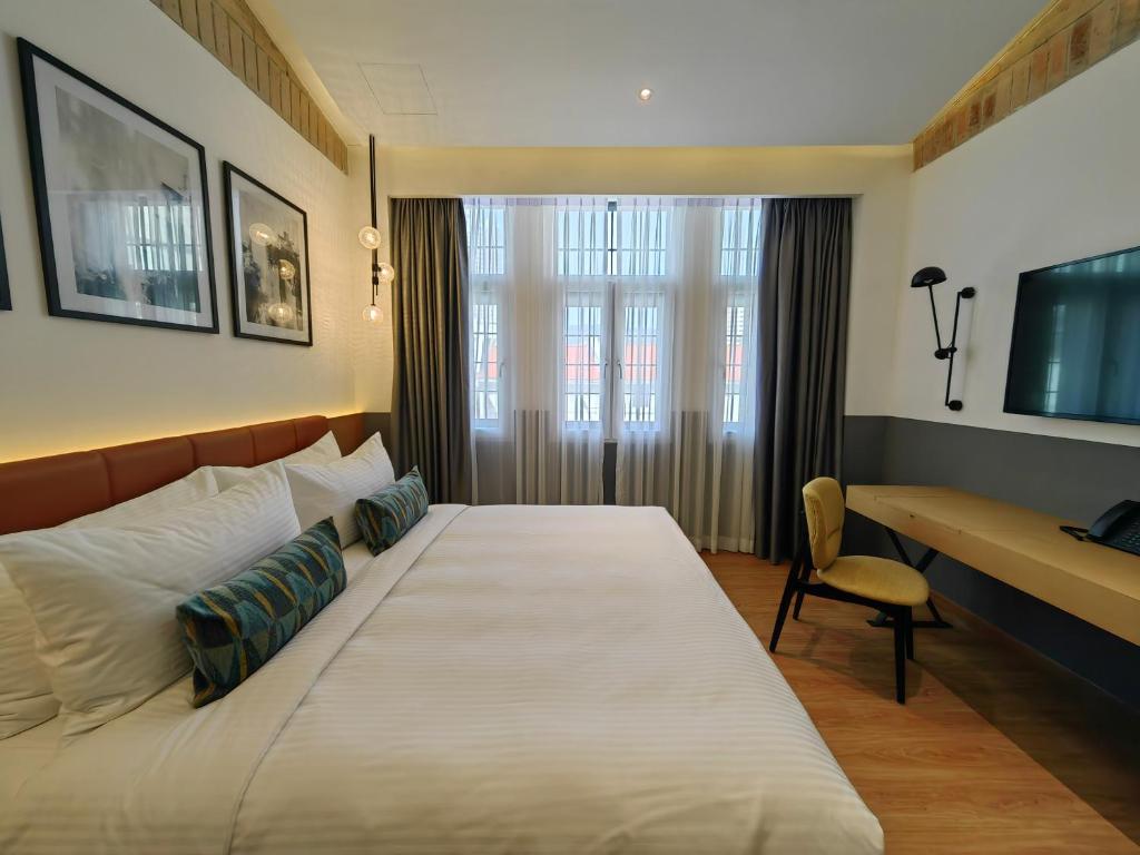 Hotel room with a large bed, white linens, and decorative teal pillows, facing a window with sheer and heavy drapes.