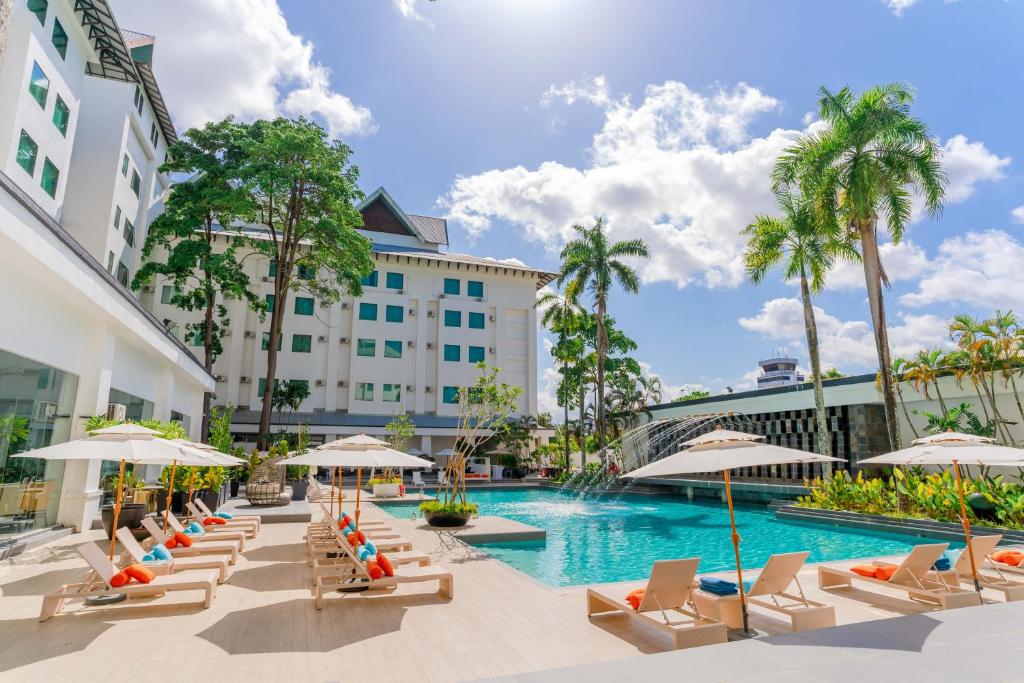 Resort pool area with white buildings, palm trees, and sun loungers under umbrellas by the water.