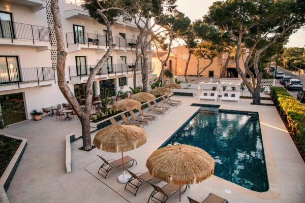 Hotel pool deck with thatched umbrellas, sun loungers, and a white building with balconies nearby.