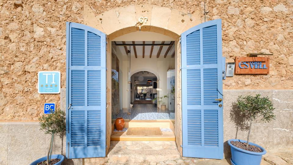 Open blue double doors set in a rustic stone wall, revealing a sunlit interior and tiled entryway with potted plants nearby.