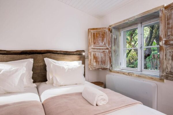 Twin bedroom with two single beds, white pillows, and a rolled towel on a light pink throw, plus a rustic window view of greenery.