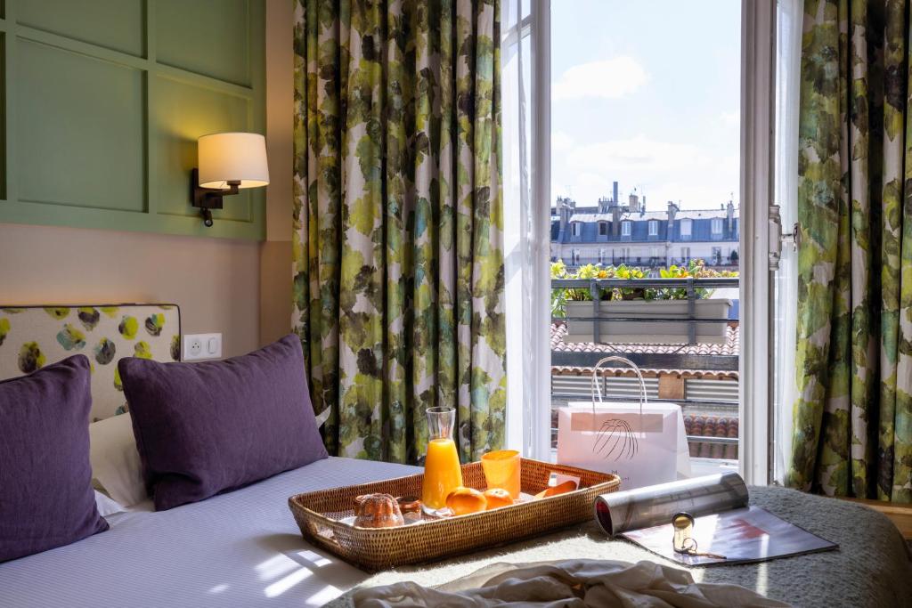 Hotel room with a breakfast tray on the bed, purple pillows, and an open balcony door overlooking city rooftops in sunlight.