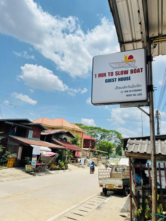 Street view with a hanging sign that reads '1 MIN TO SLOW BOAT GUEST HOUSE' along a rural road under a blue sky with scattered clouds.