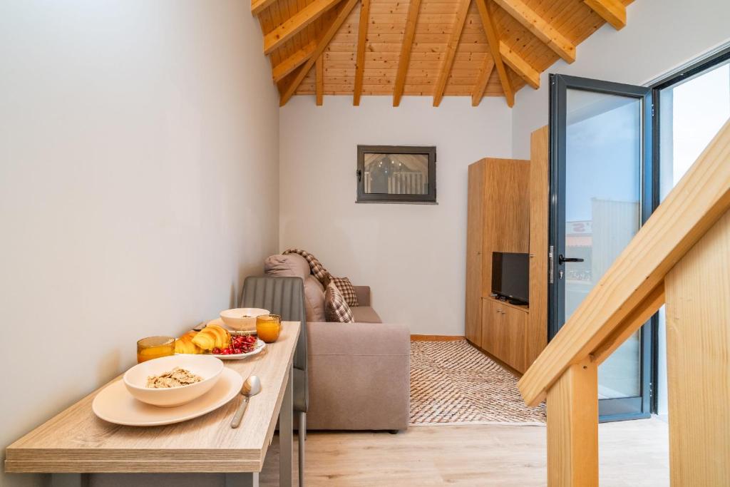Cozy loft living area with a breakfast table set with bowls, croissants, toast and orange juice, a beige sofa, and wooden vaulted ceiling beams under a large open door to the outdoors.