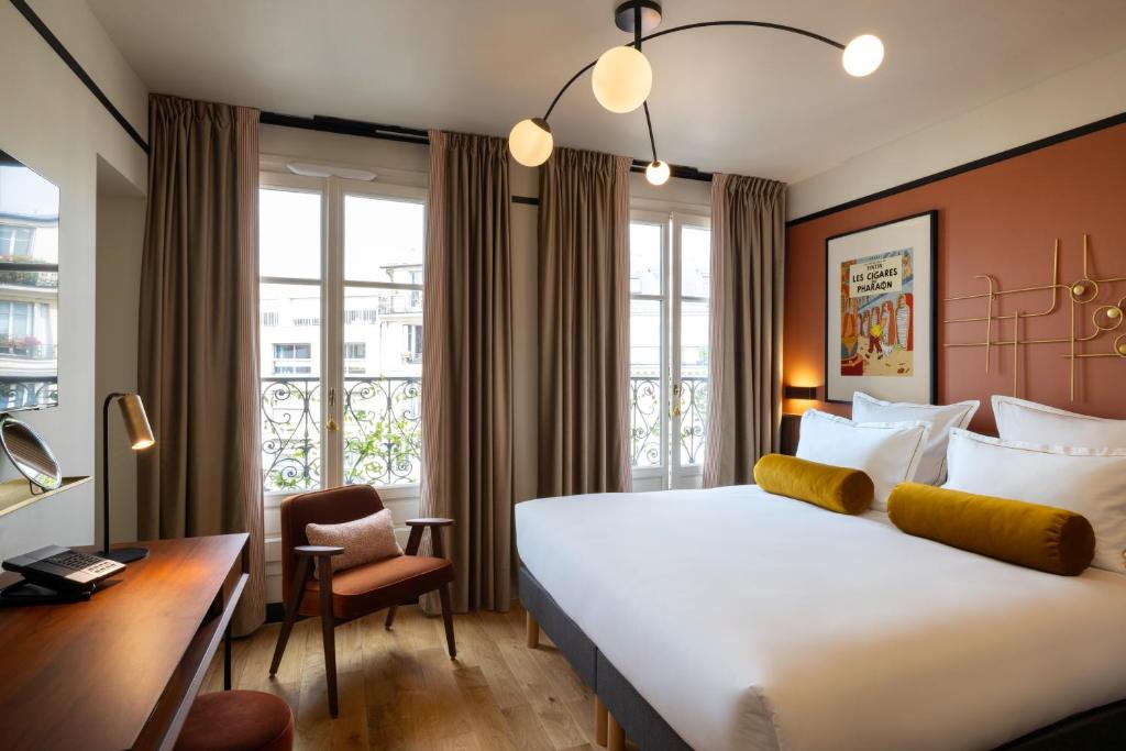 Hotel room with a large white bed and mustard bolster pillows, rust accent wall, and a desk near a windowed balcony. Framed art above the bed.