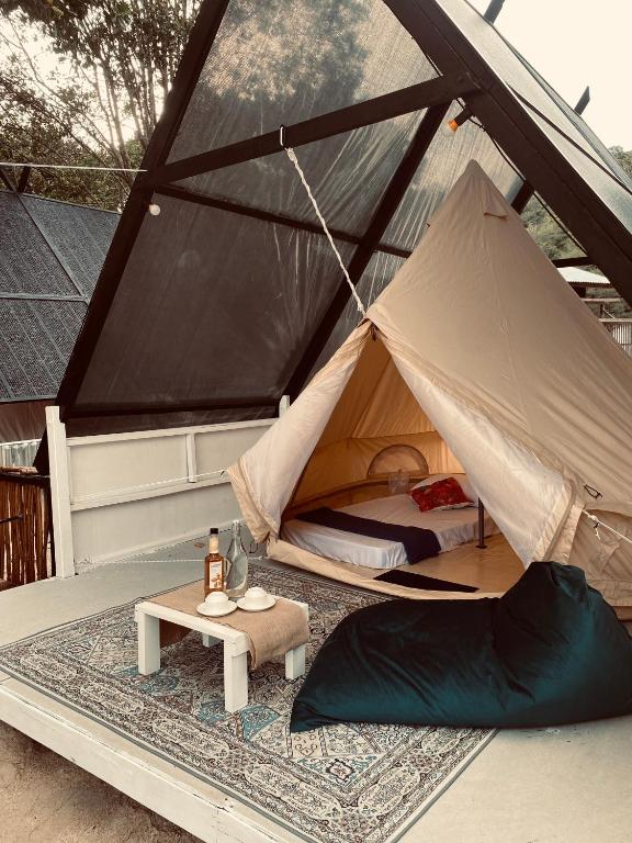 Beige rooftop tent on a deck with a bed inside, a small table with a bottle and cups, and a dark beanbag; solar panels visible nearby.
