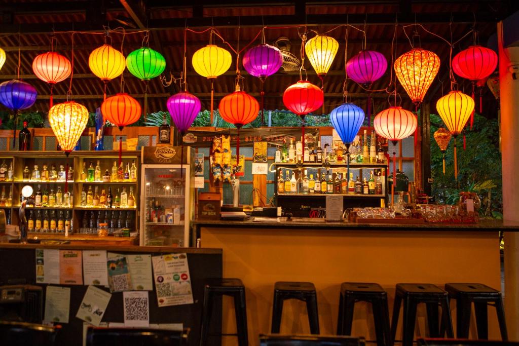 Bar interior with colorful lanterns overhead, drinks and bottles lined up behind the counter, and dark wooden stools in front.