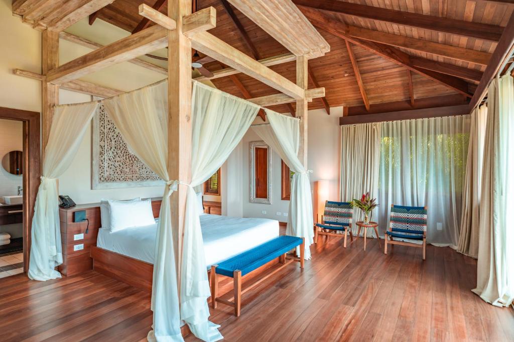 Luxurious four-poster bed with sheer drapes in a wooden-ceiling bedroom, with blue bench at foot and two patterned chairs by a windowed wall.