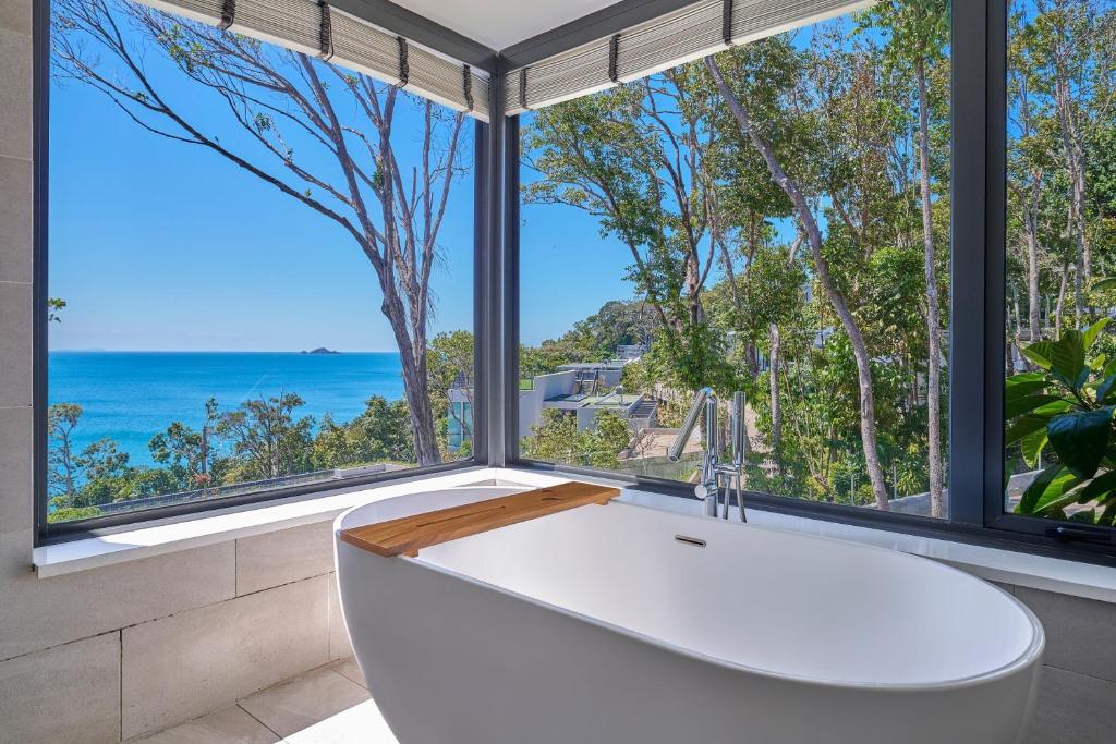 White freestanding bathtub with a wooden bath tray, set beside large windows overlooking the ocean.