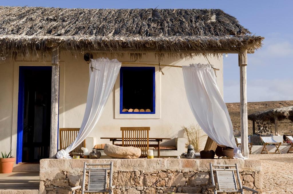 Thatched-roof beach hut with white walls, blue door/window frames, and white curtains on a sunny veranda.