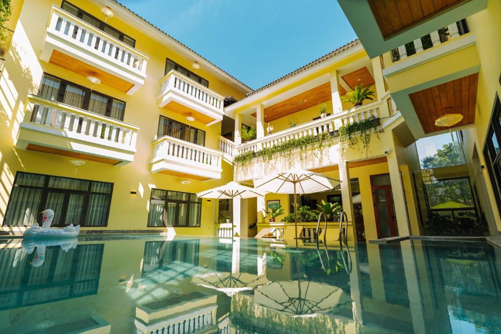 Yellow hotel courtyard with white balconies surrounding a swimming pool, blue sky above, and sun umbrellas nearby.