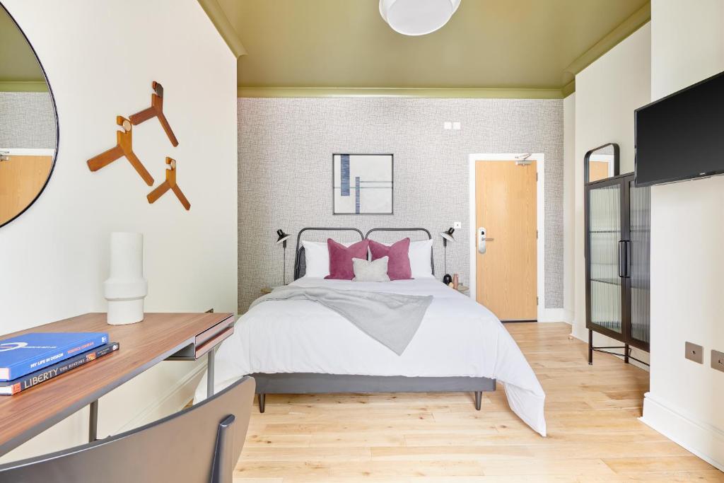 Modern hotel-style bedroom with a white bed, pink cushions, and a gray throw, plus a wooden desk and books on the left.