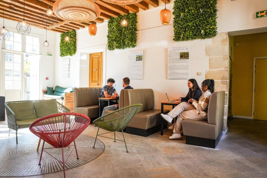 Bright lobby seating area with beige booths, hanging wicker lights, and green plant panels; three people are chatting and working on laptops nearby.