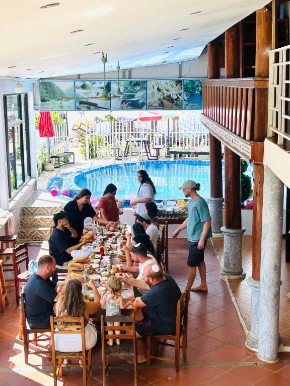 Group of people seated around a long dining table sharing a meal beside a pool in a sunny outdoor resort setting with wooden columns and murals above.