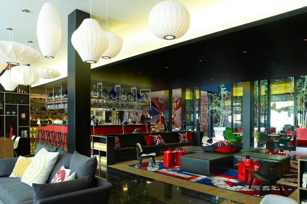 Modern hotel lobby bar with a red counter, black sofas, Union Jack cushions, white pendant lights, and glass walls.