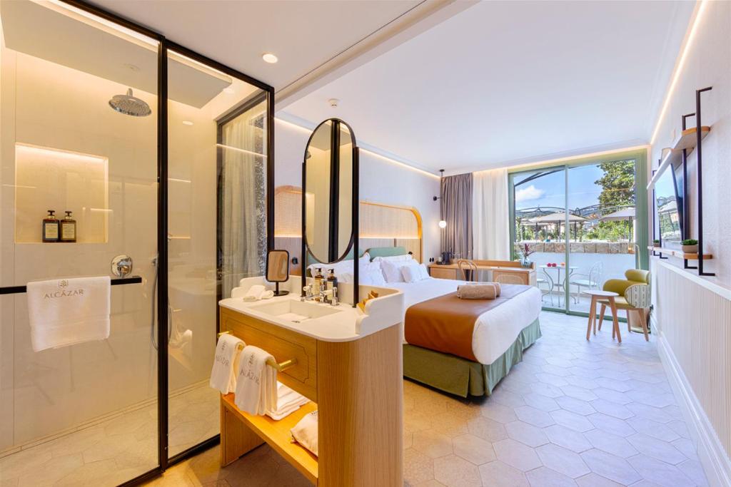 Bright hotel suite with a glass-enclosed shower, double bed, and a wooden double-sink vanity with an oval mirror.