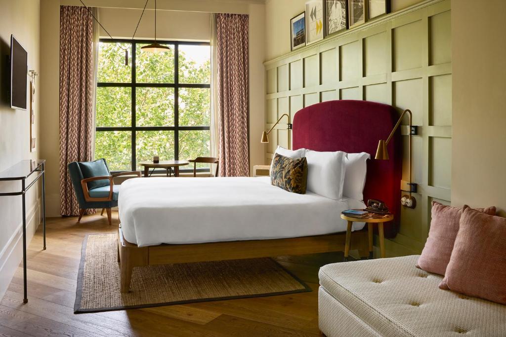Bright hotel room with a large bed and a red upholstered headboard, green paneled wall, and seating area by a big window showing trees outside.