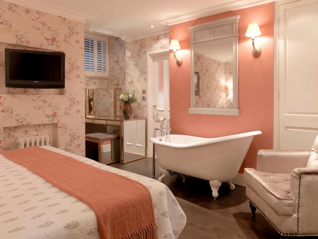 Elegant hotel room featuring a white clawfoot bathtub against a coral pink wall, floral wallpaper, wall lamps, a bed with an orange throw, and a seating chair.