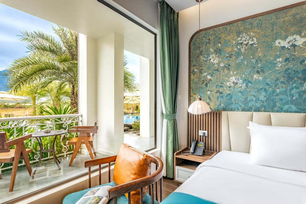 Bright hotel room with a large window opening to a balcony, palm trees outside and wooden chairs at a small table; teal floral wall behind a white bed with bedside table and lamp nearby.