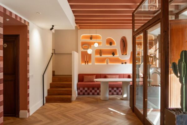 Bright, modern living/dining area with a white round table, red cushioned bench seating, warm orange backlit shelves, staircase on the left, and glass doors on the right with a tall cactus nearby.