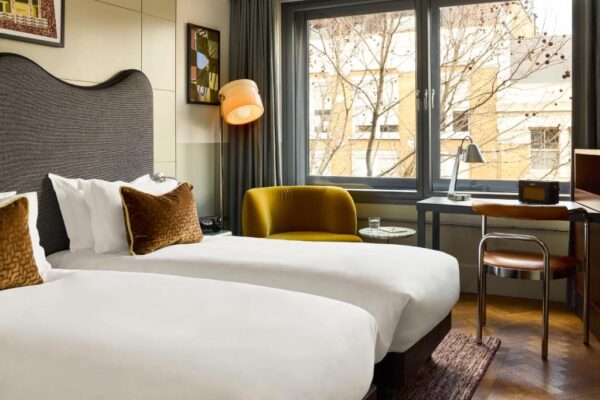 Hotel room with two white-duvet beds, brown accent pillows, a mustard chair, and a desk by a large window overlooking a tree-lined street.