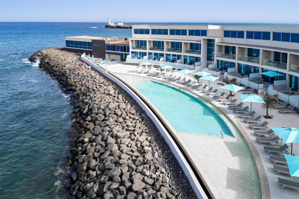 Modern seaside hotel with a curved turquoise pool, loungers, and blue umbrellas along the terrace by the ocean.