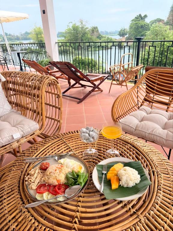 Outdoor wicker patio with a dining table set, two plates of food, fruit, and a glass of juice, overlooking a lake and greenery.