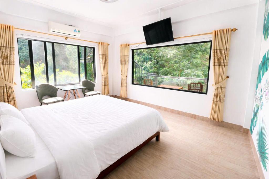 Bright hotel room with a large bed, beige curtains, and a balcony view of greenery beyond the windows; wall-mounted TV nearby.