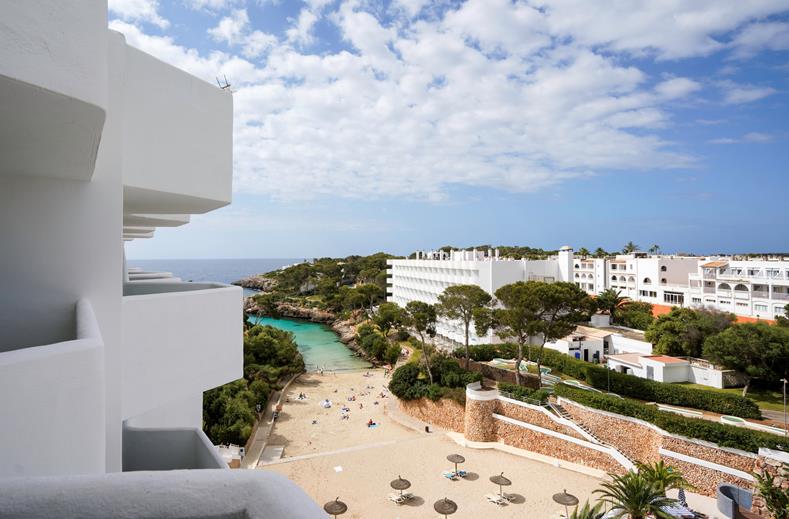 White resort buildings overlook a sandy beach with a turquoise cove and scattered sun umbrellas and loungers by the water.