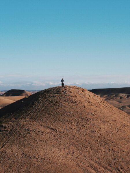 Person standing on top of a sandy hill in a desert under a clear blue sky.